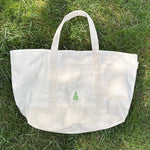 A natural canvas tote bag with two top handles and a small green tree logo on the front, placed on a green grass lawn.