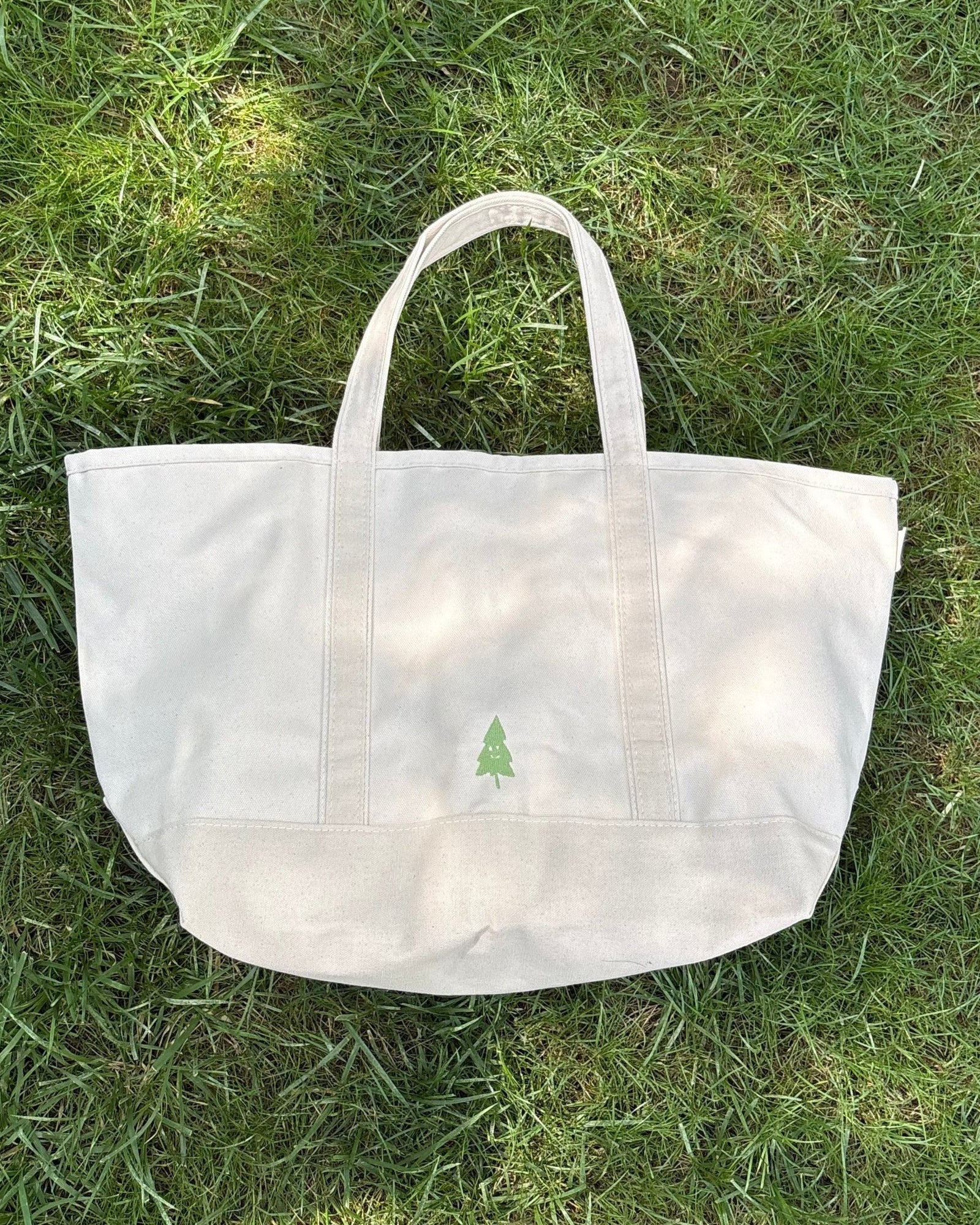 A natural canvas tote bag with two top handles and a small green tree logo on the front, placed on a green grass lawn.