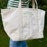An off-white canvas tote bag with green embroidered text reading 'this bag has seen things' on the front pocket, carried by a person wearing a green and white striped sweater and blue jeans in an outdoor garden setting.
