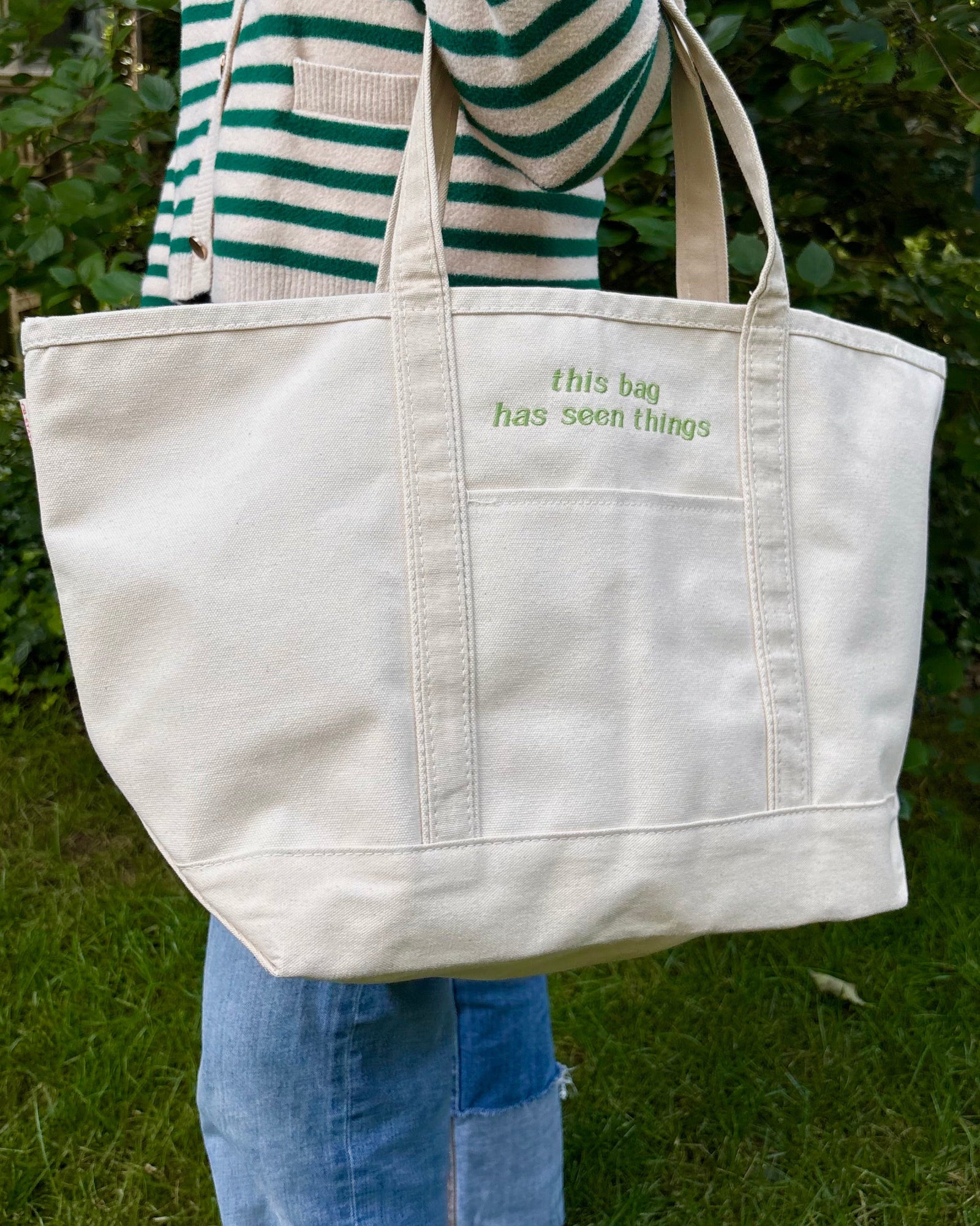 An off-white canvas tote bag with green embroidered text reading 'this bag has seen things' on the front pocket, carried by a person wearing a green and white striped sweater and blue jeans in an outdoor garden setting.