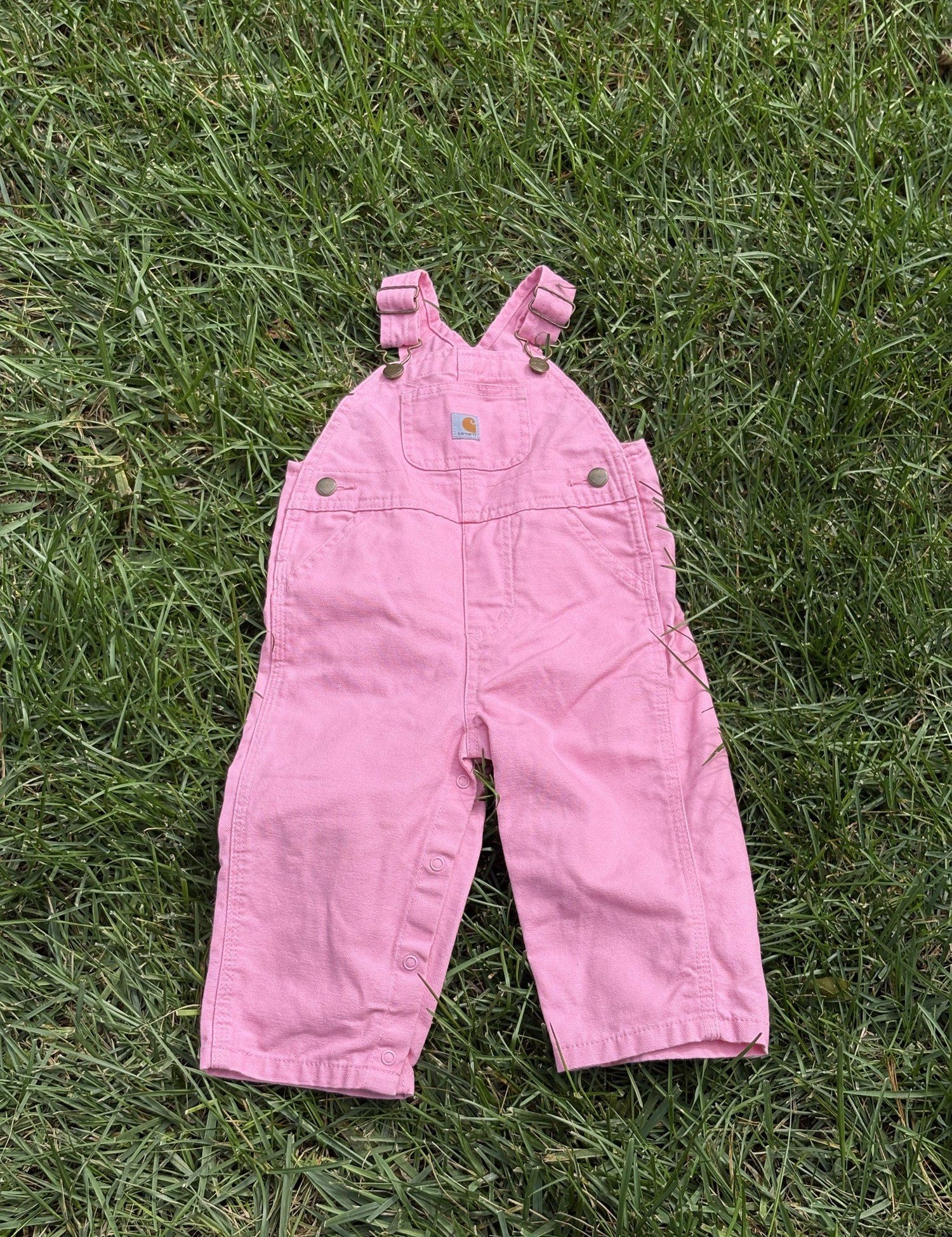 Pre-loved Carhartt pink canvas bib overalls, kids size 12M, back view, laid flat on grass