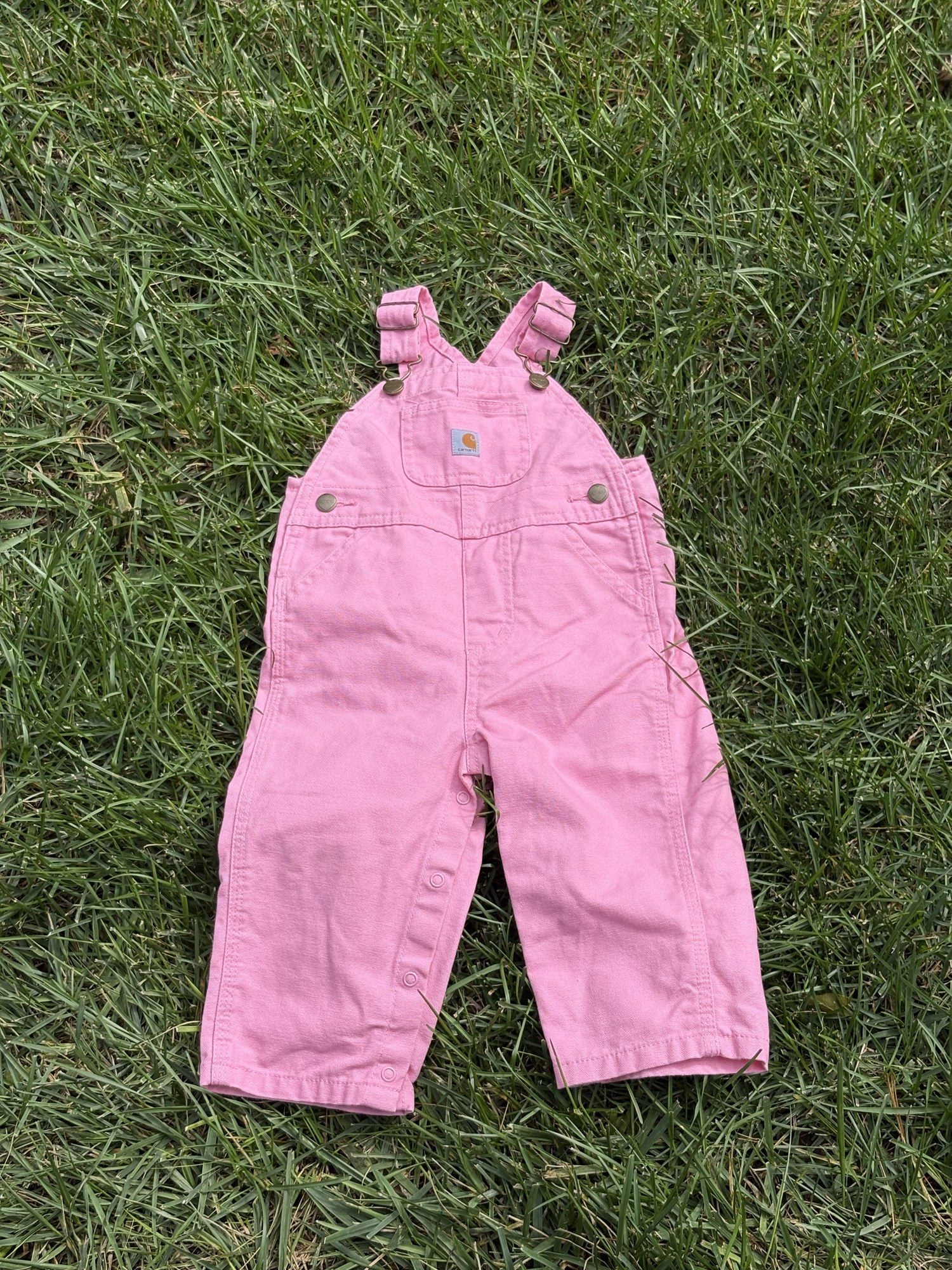 Pre-loved Carhartt pink canvas bib overalls, kids size 12M, back view, laid flat on grass