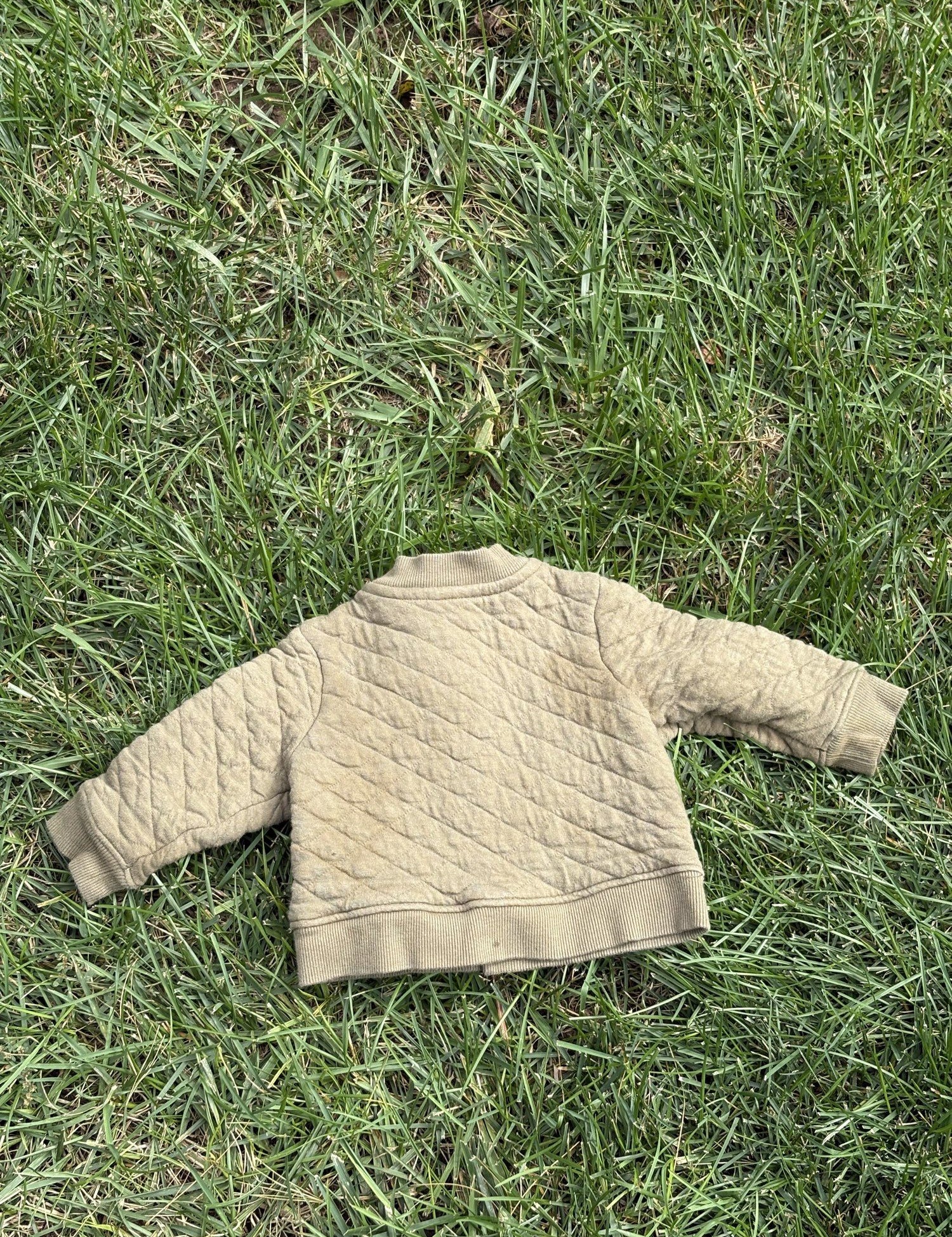 Pre-loved Hanna Andersson khaki quilted snap-front jacket, kids size 6-12M, back view, laid flat on grass