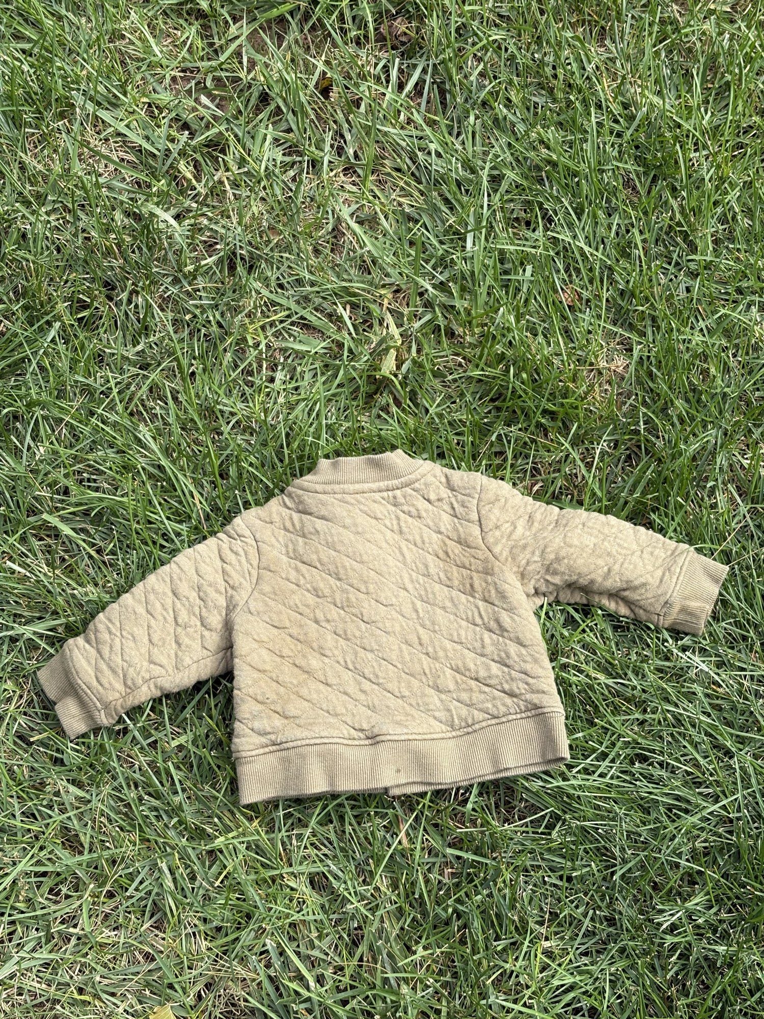 Pre-loved Hanna Andersson khaki quilted snap-front jacket, kids size 6-12M, back view, laid flat on grass