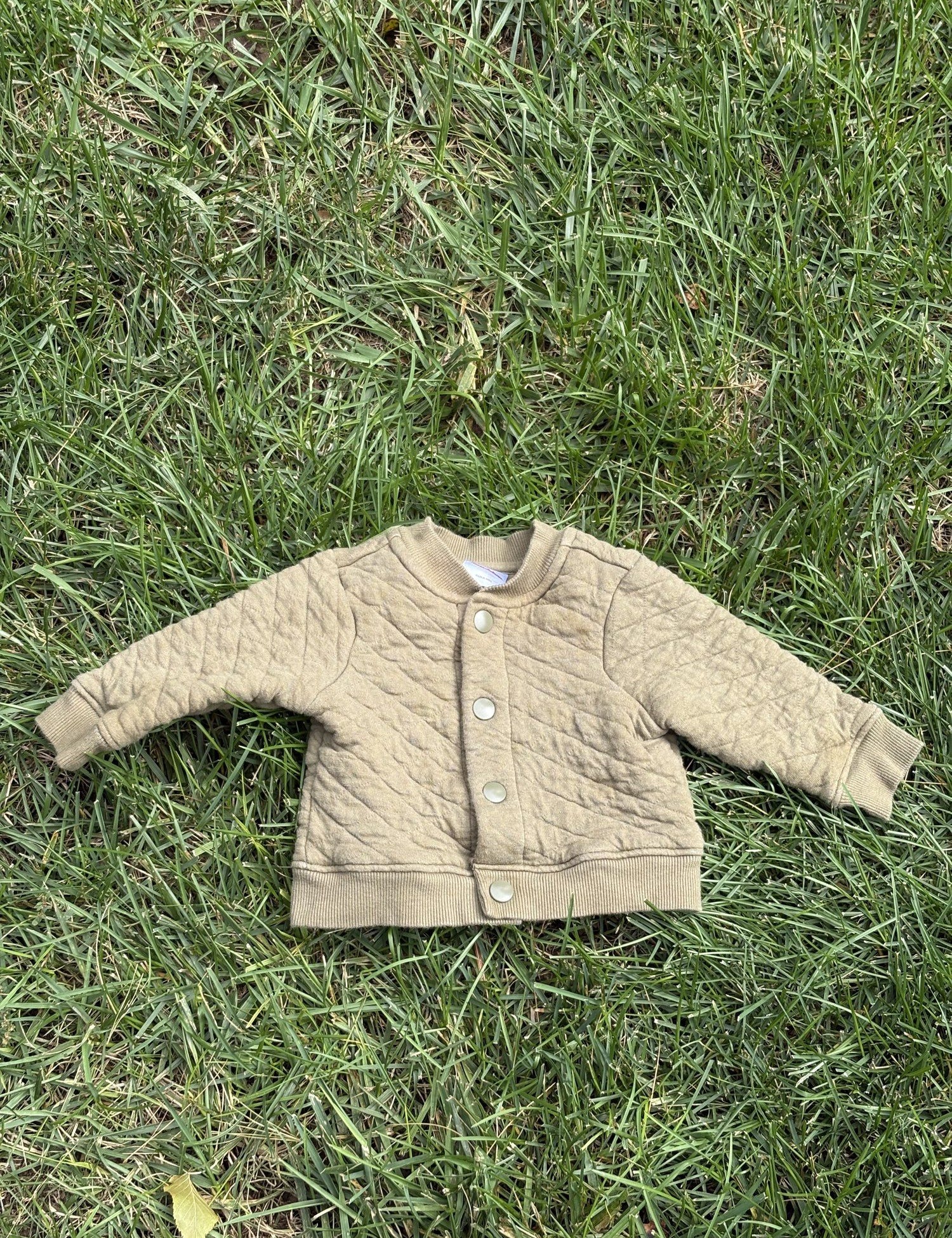 Pre-loved Hanna Andersson khaki quilted snap-front jacket, kids size 6-12M, front view, laid flat on grass