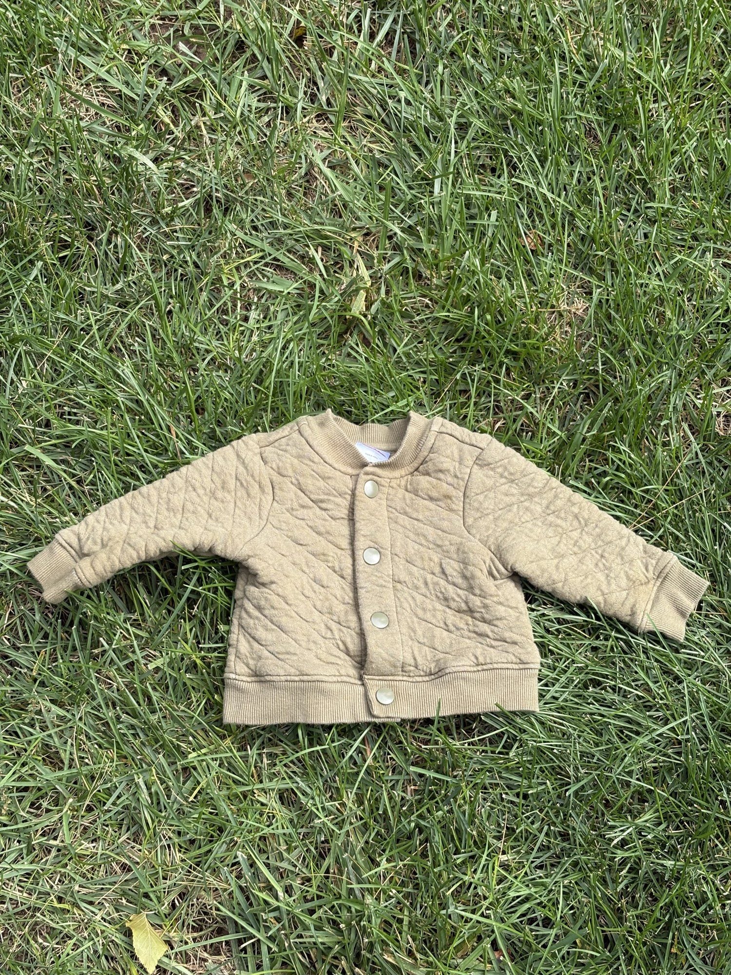 Pre-loved Hanna Andersson khaki quilted snap-front jacket, kids size 6-12M, front view, laid flat on grass