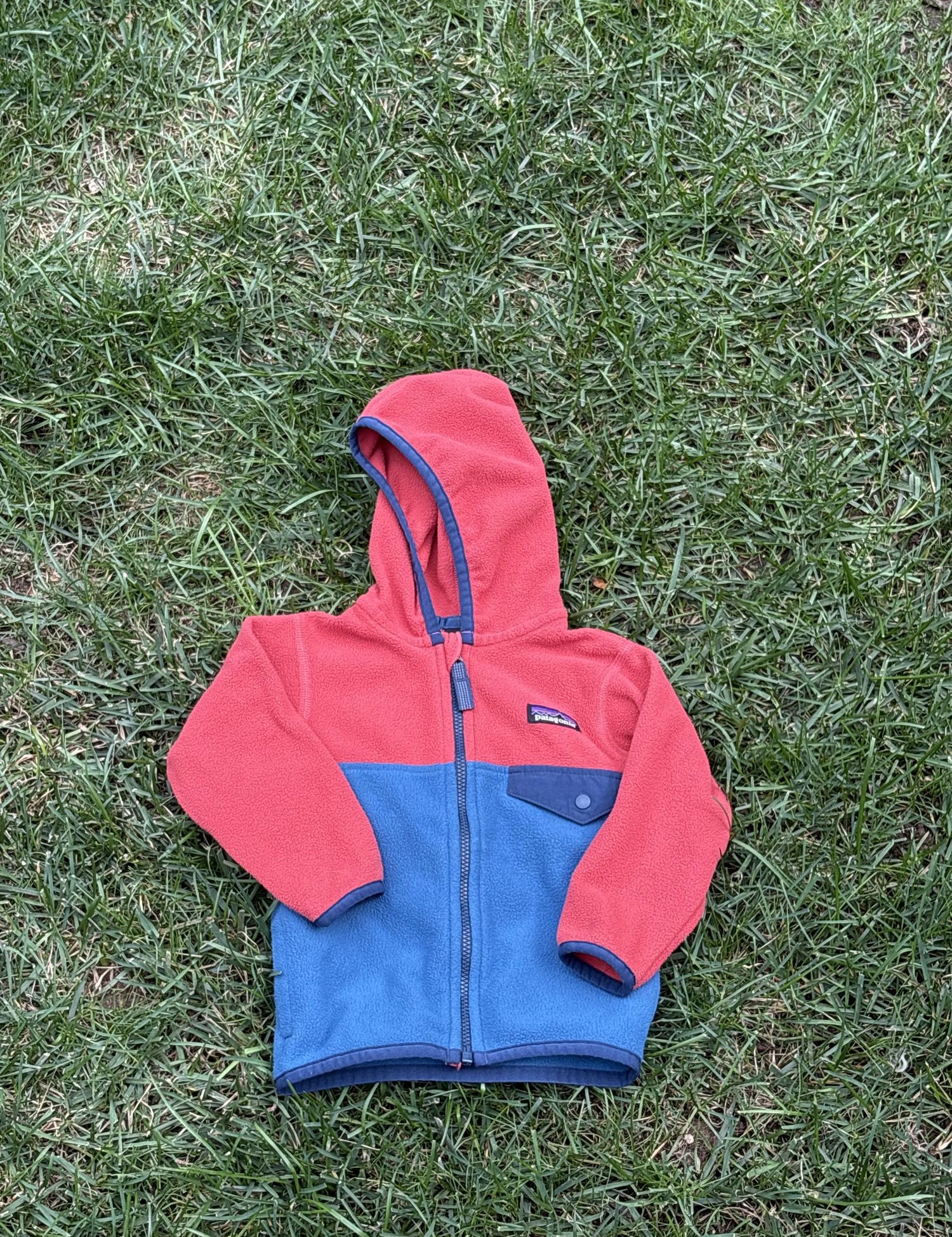 Pre-loved Patagonia red and blue colorblock hooded fleece jacket, kids size 6-12M, front view, laid flat on grass
